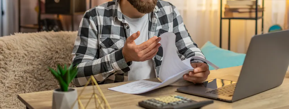 Homem lê documentos sobre a mesa de casa, ao lado de uma calculadora e de um notebook